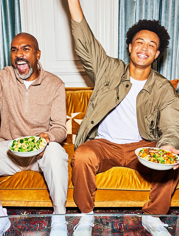 Two men laughing on a couch, holding bowls of Daily Harvest meals