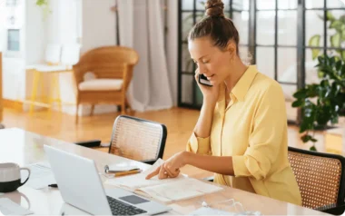 Woman in a yellow shirt talking on the phone, pointing at a notebook, with a laptop and coffee cup on the table.