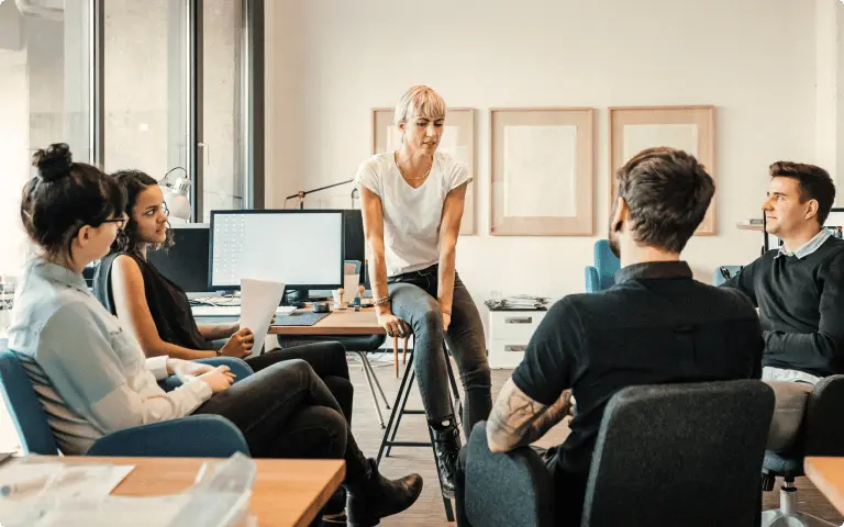 A group of five people having a discussion in an office setting, with one person sitting on a desk and others seated around.