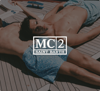 Zwei Männer in Badehosen liegen auf einem Boot. Über dem Bild steht das Logo "MC2 Saint Barth".