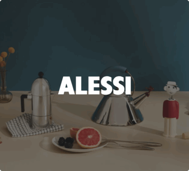 Utensilios de cocina de Alessi sobre una mesa: una tetera, una cafetera y un plato de fruta. La palabra «ALESSI» se muestra de manera prominente.
