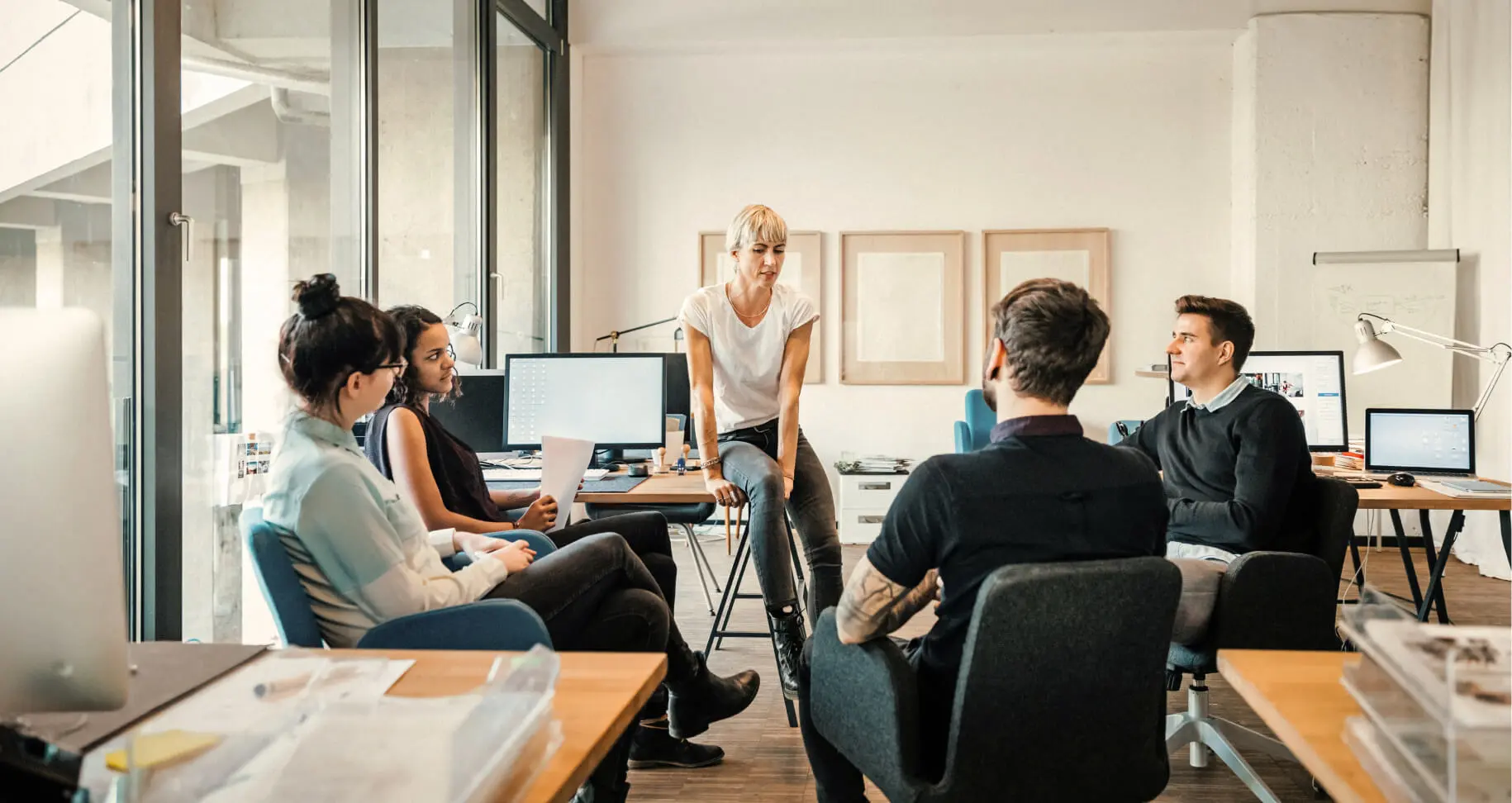Un grupo de cinco personas reunidas en una oficina moderna con ordenadores y ventanas grandes.