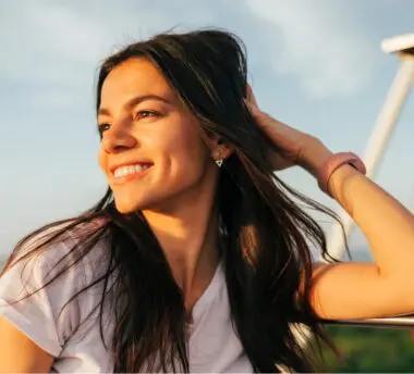 A young brunette sits outside teasing her hair with her hand. She smiles, her sunlight face glowing.