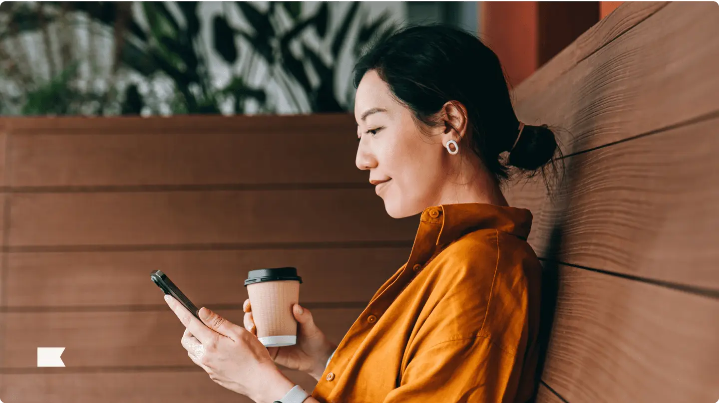 Woman in an orange shirt holding a coffee cup and looking at her phone, sitting against a wooden wall.