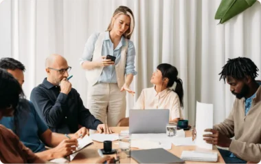 A group of people in a meeting room, with one person standing and speaking, while others sit and listen, taking notes.