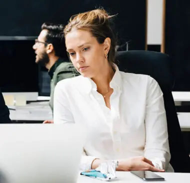 A woman working on her laptop with others in the office in the background