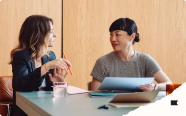 Two women sitting at a table, engaged in conversation, with documents and a glass of water in front of them.