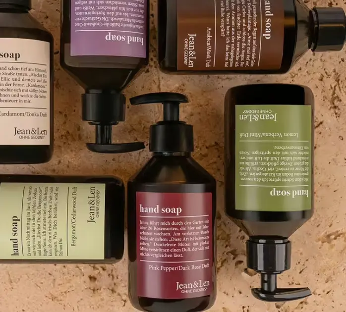 Bottles of Jean & Len hand soap with various labels and scents, arranged on a textured surface.