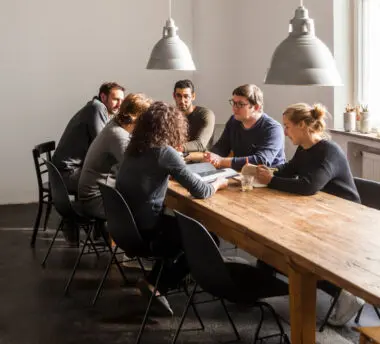 Coworkers in a modern office gather at a long wooden table, talking and flipping through books