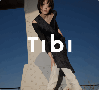 Person in a stylish black outfit and white pants poses against a metal structure. "Tibi" text overlays the image.