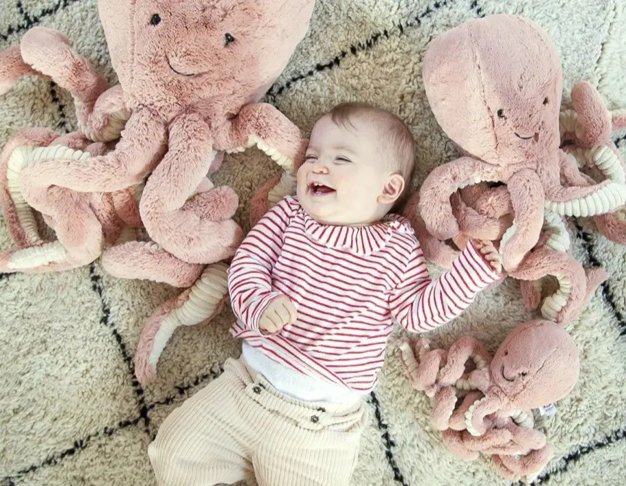 A smiling baby lying on the floor amongst several pink stuffed octopuses.