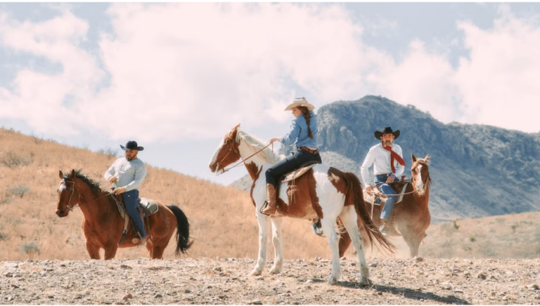 Three people are wearing Tecovas brand cowboy hats and riding horses. There are mountains in the background.