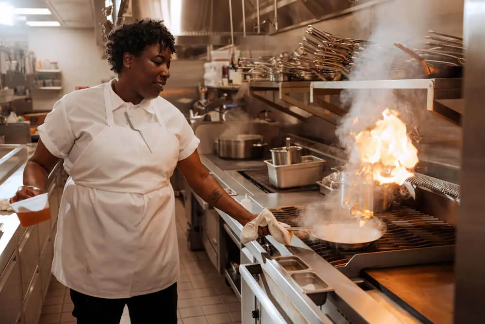 Image shows a chef in a busy restaurant kitchen, flipping something on fire in a pan on the stove.