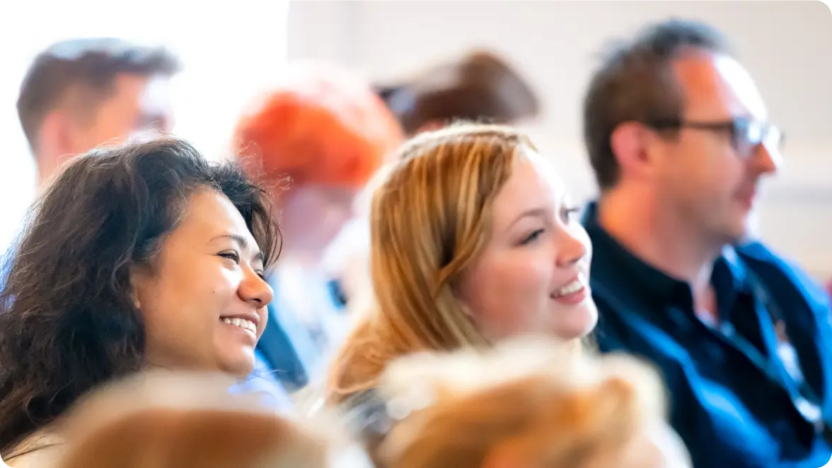 Audience members smile and engage during a live event or presentation, with soft lighting and a warmly lit background creating a welcoming atmosphere.