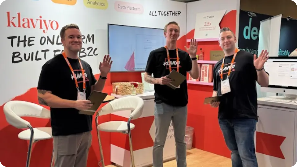 Three smiling Klaviyo team members wave at the camera while standing in front of a trade show booth. The backdrop reads "The only CRM built for B2C" and features product categories like Email, Analytics, and Data Platform.