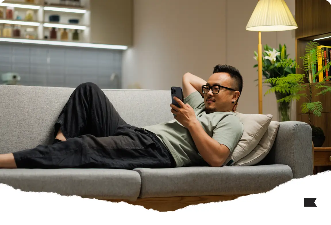 Man relaxing on a couch, looking at a smartphone. There's a lamp and plants in the background.