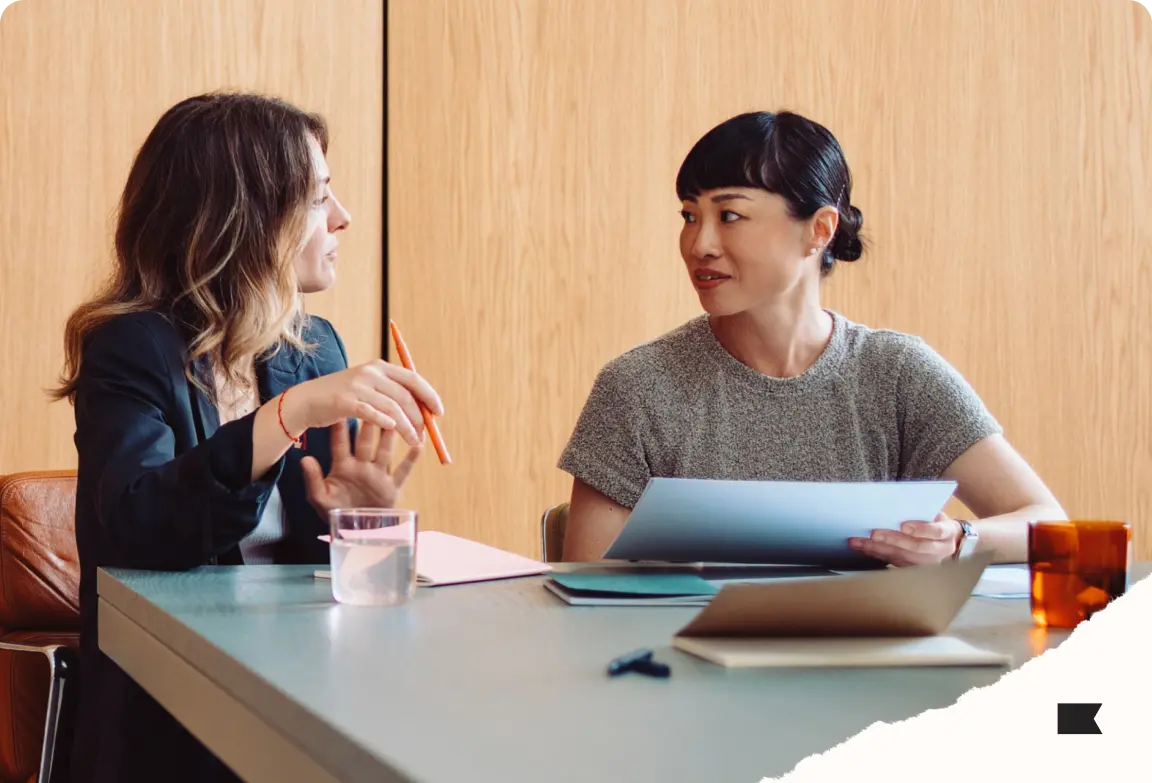 Two women sitting at a table, engaged in conversation. One holds a pen, the other holds documents. Office setting.