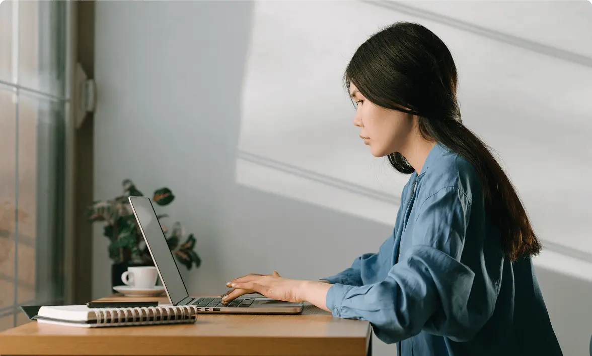 Woman in a blue shirt working on a laptop at a desk with a notebook and coffee cup nearby.
