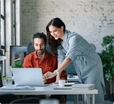 Two colleagues collaborating at a desk with a laptop, one seated and one standing, in a modern office setting.