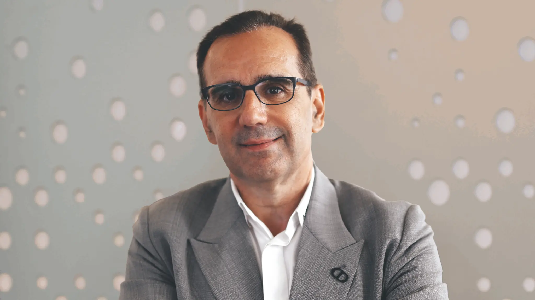 Man in a gray suit and glasses, smiling slightly, with a blurred background featuring circular patterns.